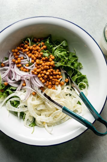 An overhead shot shows a large bowl with baby arugula, shaved red onion, and shaved fennel, and crispy air fried chickpeas. A set of tongs also sticks out of the bowl.