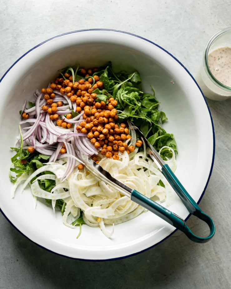 An overhead shot shows a large bowl with baby arugula, shaved red onion, and shaved fennel, and crispy air fried chickpeas. A set of tongs also sticks out of the bowl.