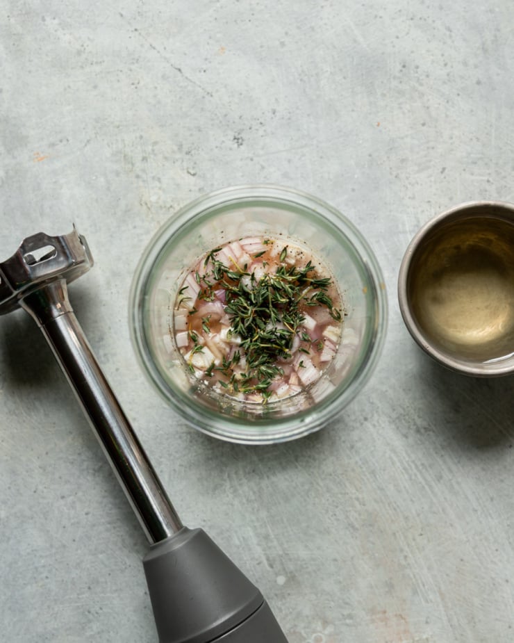 An overhead shot shows a jar filled with ingredients for a shallot thyme dressing. To the side, you see a small bowl of oil and an immersion blender.
