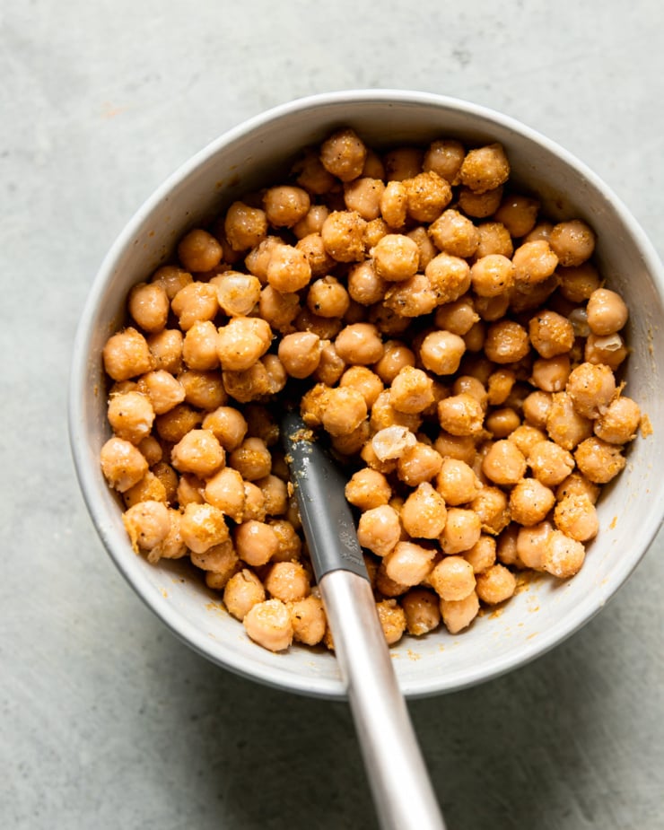 An overhead shot shows a bowl of chickpeas tossed with oil, salt, pepper, nutritional yeast, onion powder, and garlic powder. A spatula is sticking out of the bowl.