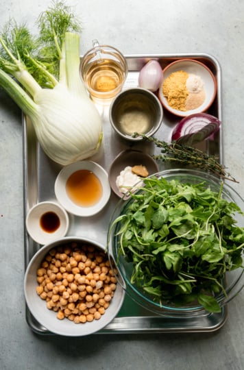 An overhead shot shows ingredients needed for a chickpea arugula salad; all sitting on a metal sheet pan.