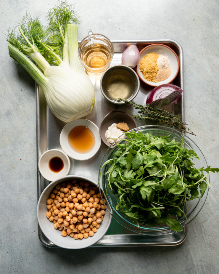 An overhead shot shows ingredients needed for a chickpea arugula salad; all sitting on a metal sheet pan.