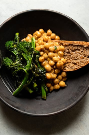A dark ceramic bowl containing mustard-braised chickpeas, roasted broccoli rabe with charred edges, and a thick slice of whole grain sourdough bread. A fork rests on the left side of the bowl. Shot from overhead on a light gray surface.
