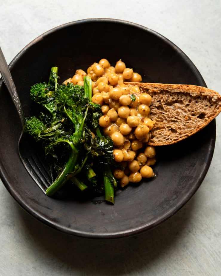 A dark ceramic bowl containing mustard-braised chickpeas, roasted broccoli rabe with charred edges, and a thick slice of whole grain sourdough bread. A fork rests on the left side of the bowl. Shot from overhead on a light gray surface.