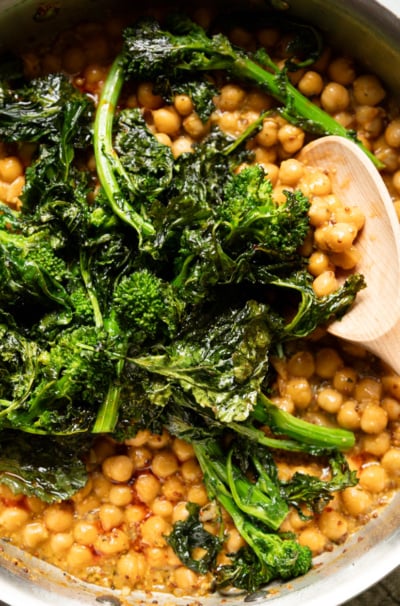 Close-up overhead shot of a stainless steel skillet filled with golden mustard braised chickpeas in a glossy sauce, topped with a large mound of wilted, charred broccoli rabe. A wooden spoon rests in the pan on the right side.