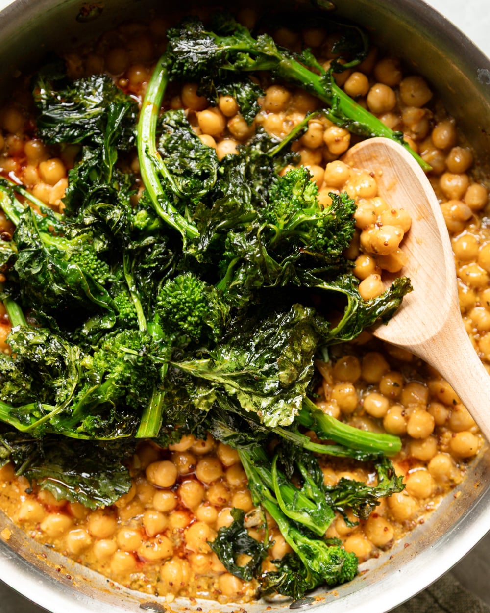 Close-up overhead shot of a stainless steel skillet filled with golden mustard braised chickpeas in a glossy sauce, topped with a large mound of wilted, charred broccoli rabe. A wooden spoon rests in the pan on the right side.