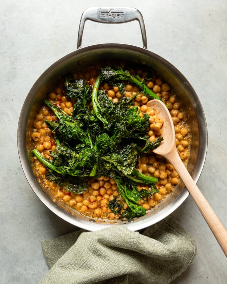 Overhead view of an All-Clad stainless steel skillet on a light gray surface, containing braised chickpeas in a saucy broth with a pile of roasted broccoli rabe in the center. A wooden spoon leans across the right side of the pan, and a folded green linen napkin sits beneath the pan's handle.