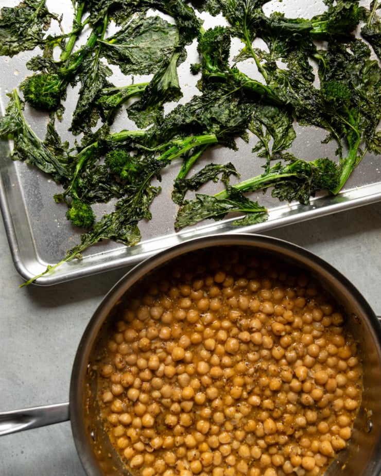 Two vessels side by side on a gray surface: a rimmed metal baking sheet in the upper half holding roasted, slightly charred broccoli rabe, and a stainless steel skillet in the lower half filled with golden chickpeas in a glossy braising liquid.