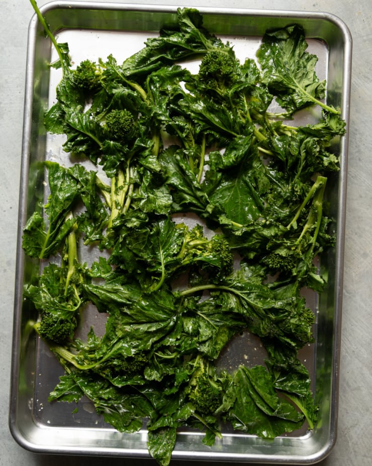 Overhead shot of a rimmed metal baking sheet covered with raw broccoli rabe — leafy, bright green stalks with small florets — laid out flat and glistening with oil, ready to roast.