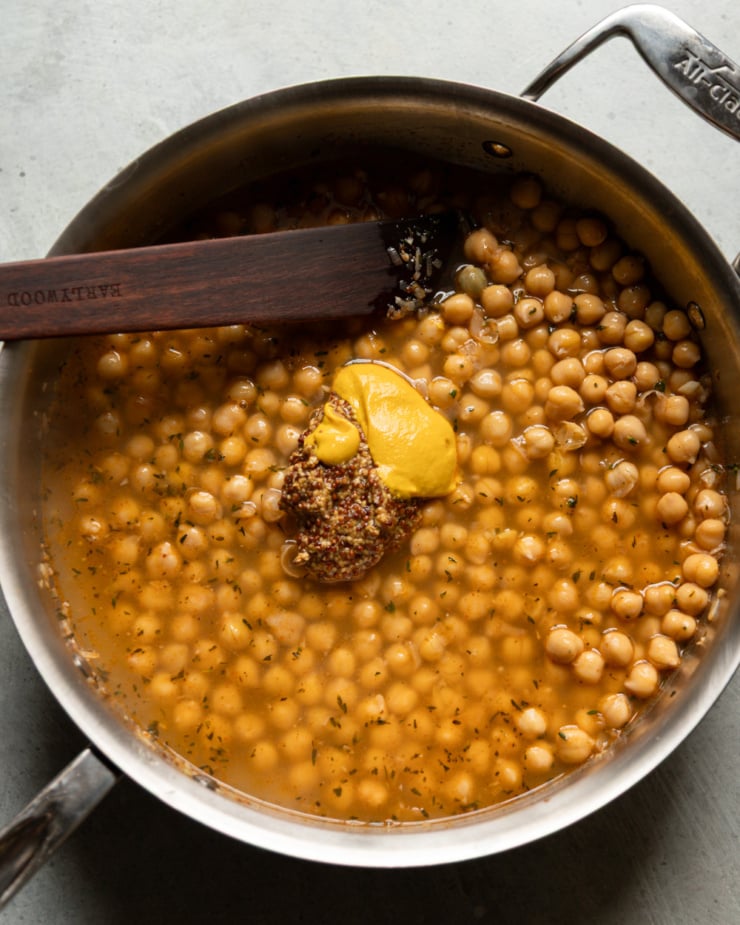 Close-up overhead view of an All-Clad saucepan with chickpeas simmering in a golden broth. A large dollop of smooth yellow mustard and a spoonful of whole grain mustard have just been added to the pan, sitting on top of the chickpeas. A dark wooden spatula rests in the pan.