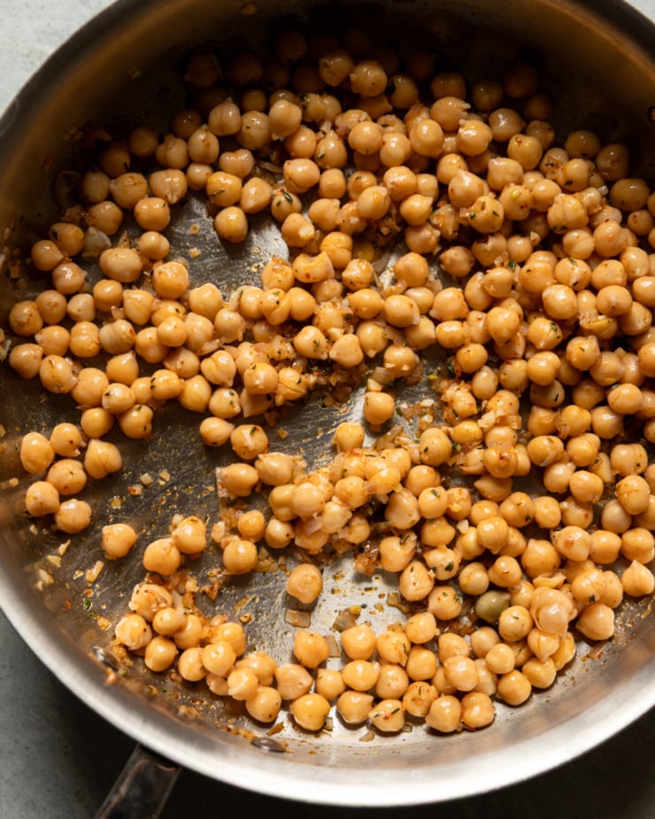 Overhead close-up of chickpeas toasting in a dry stainless steel skillet. The chickpeas are golden and scattered across the pan, coated in minced shallots, garlic, and thyme, with some browning visible on the pan surface.