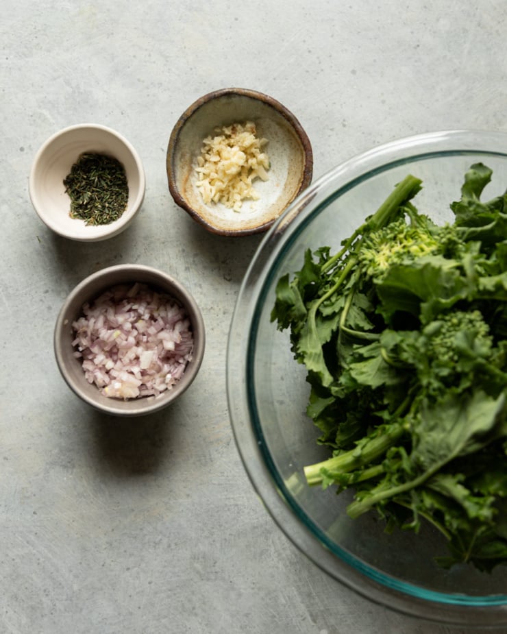 Flat lay of prepped ingredients on a gray surface: a large glass bowl of fresh broccoli rabe on the right, and three small ceramic bowls arranged to the left containing minced garlic, diced shallots, and minced thyme respectively.