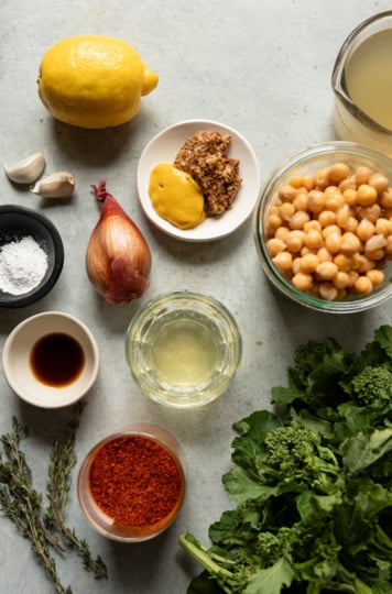 Flat lay of all recipe ingredients arranged on a gray surface: a whole lemon, two garlic cloves, a shallot, a small bowl of yellow and whole grain mustard, a jar of chickpeas, a measuring cup of vegetable broth, a bowl of ground chilies, a small bowl of white wine, a dish of vegan Worcestershire, a bowl of arrowroot powder, fresh thyme sprigs, and a bunch of fresh broccoli rabe