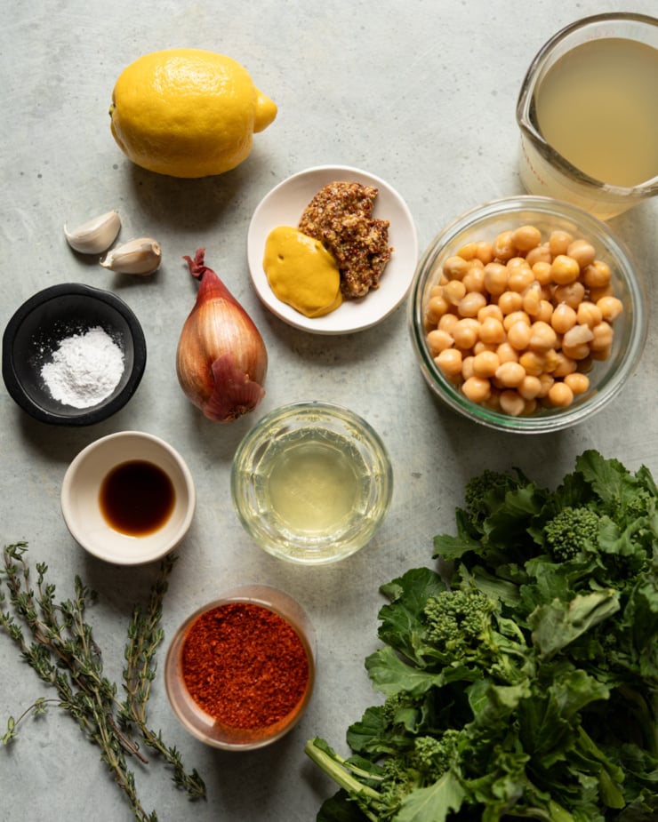 Flat lay of all recipe ingredients arranged on a gray surface: a whole lemon, two garlic cloves, a shallot, a small bowl of yellow and whole grain mustard, a jar of chickpeas, a measuring cup of vegetable broth, a bowl of ground chilies, a small bowl of white wine, a dish of vegan Worcestershire, a bowl of arrowroot powder, fresh thyme sprigs, and a bunch of fresh broccoli rabe