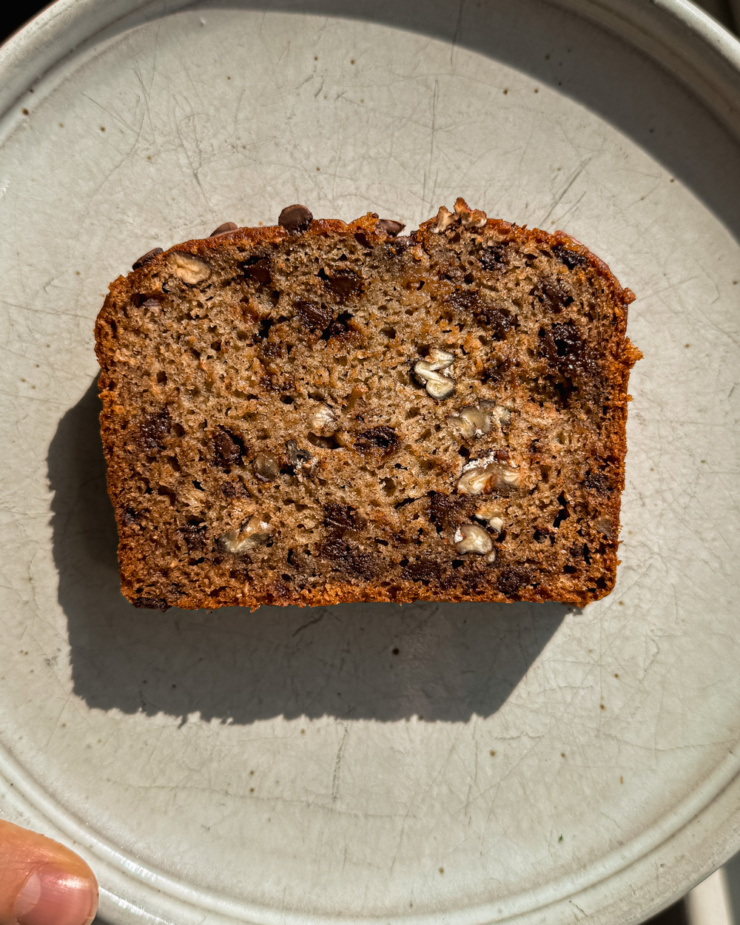 An up close, overhead shot shows a slice of vegan banana bread on a plate with lots of chocolate chips and morsels of chopped pecan as well.