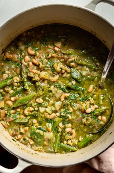 An overhead shot shows a Dutch oven filled with spring green minestrone soup. A ladle is sticking out of the soup to the side.