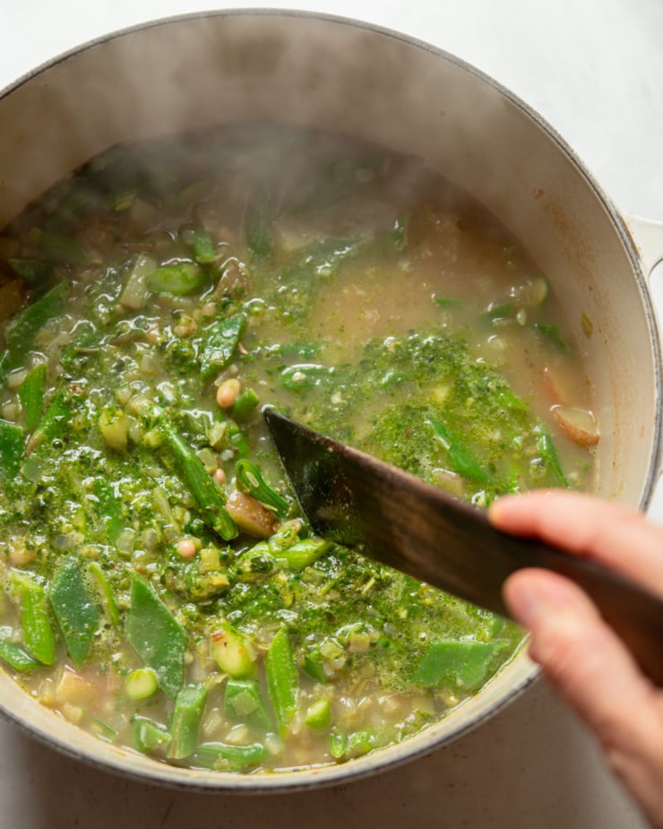 An overhead shot shows a hand using a wooden utensil to stir pesto into a mixed vegetable, white bean, and pasta soup.