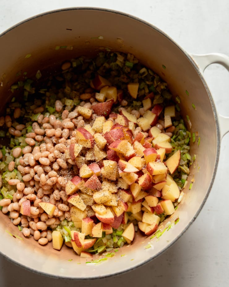 An overhead shot shows a Dutch oven pot with sautéed aromatic vegetables, white beans, and chopped potatoes inside.