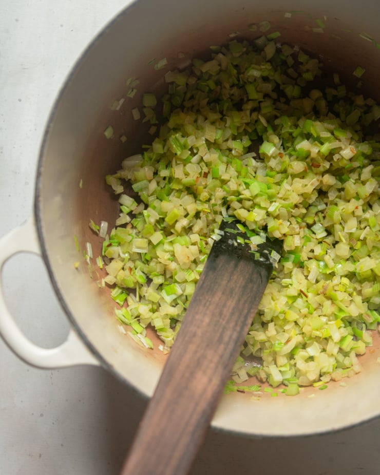 An overhead shot shows a sautéed mixture of celery, onions, fennel, and leeks in a Dutch oven pot. A wooden stirring utensil is sticking out of the pot.