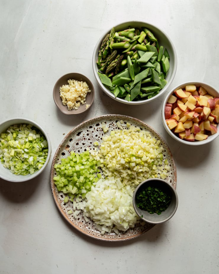 An overhead shot shows bowls and a plate filled with the following: chopped asparagus and green beans, chopped potatoes, chopped parsley, chopped onions, chopped celery, chopped fennel, chopped leeks and minced garlic.