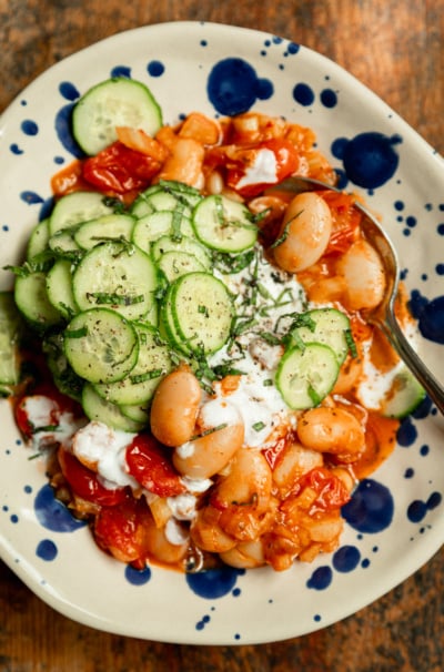 Overhead shot of a finished bowl of harissa butter beans topped with sliced cucumber mint salad and dollops of vegan yogurt, served on a blue-speckled ceramic plate with a spoon.