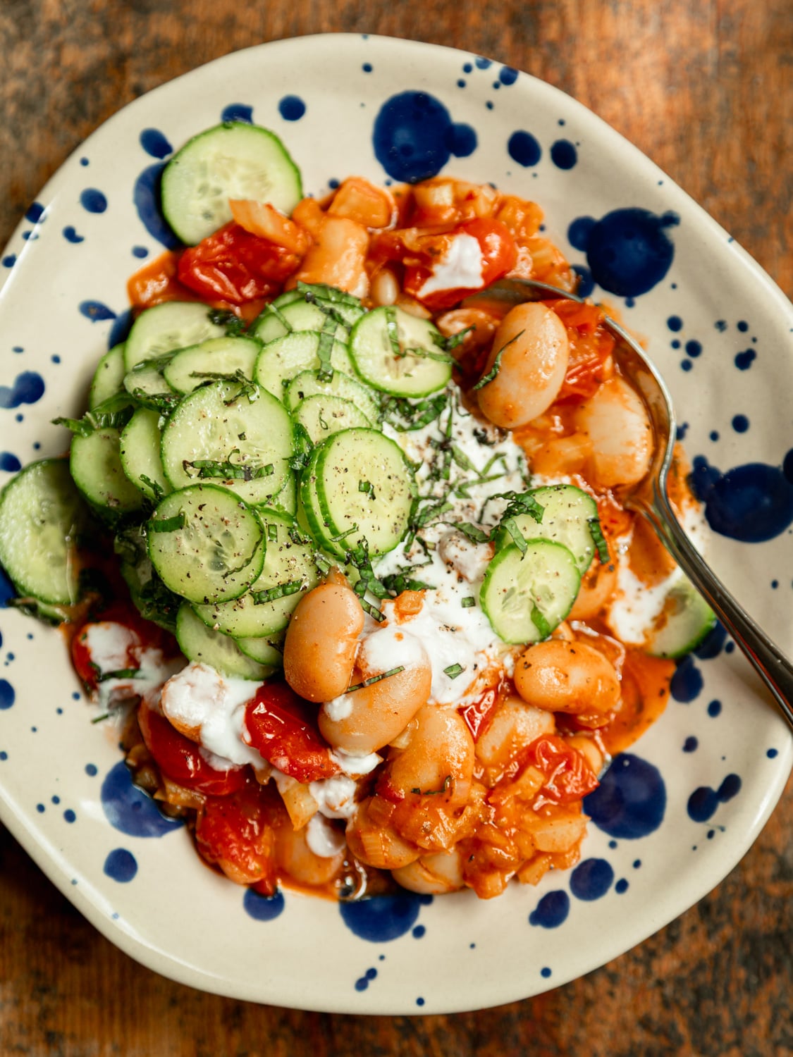 Overhead shot of a finished bowl of harissa butter beans topped with sliced cucumber mint salad and dollops of vegan yogurt, served on a blue-speckled ceramic plate with a spoon.