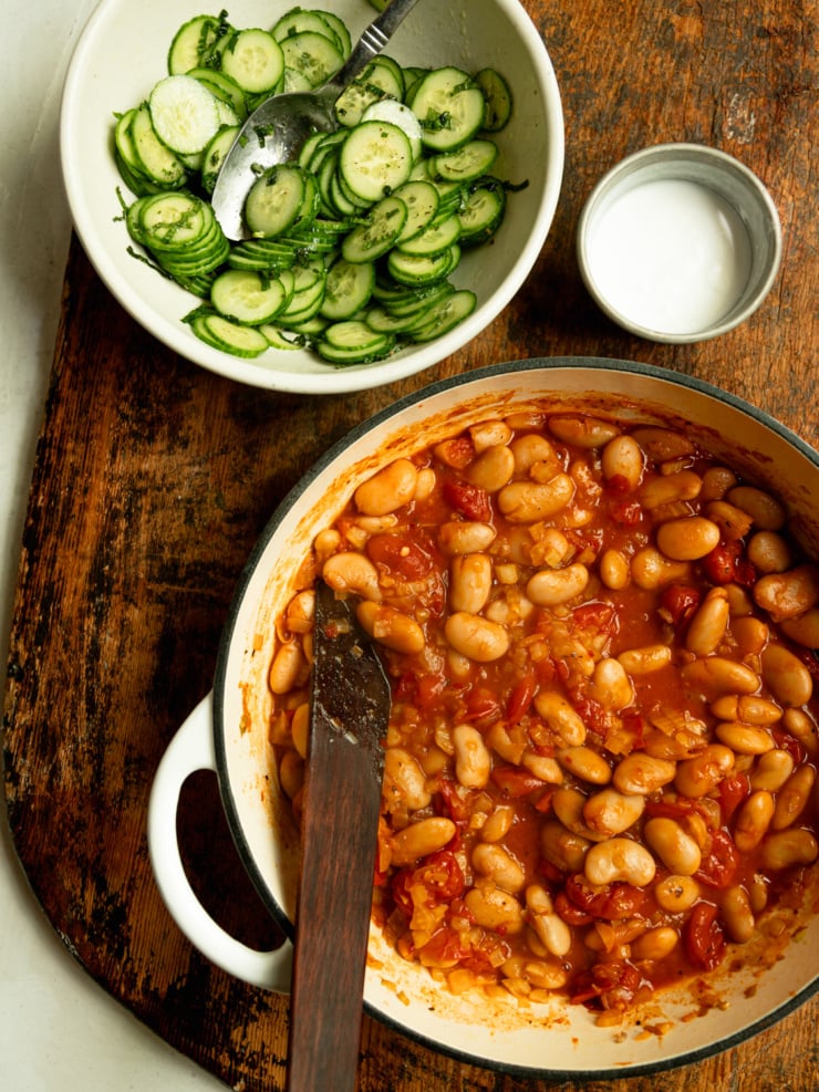 Overhead shot of the completed dish components: a Dutch oven filled with harissa butter beans in tomato sauce, a bowl of sliced cucumber mint salad, and a small tin of vegan yogurt, arranged on a dark wooden board.