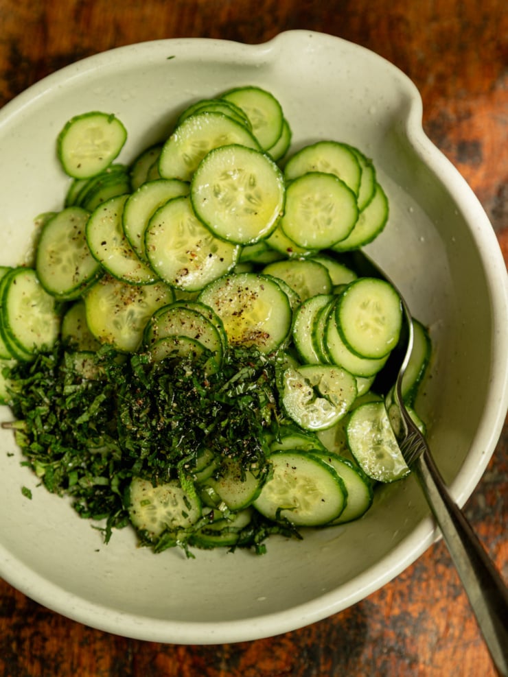 Close-up overhead shot of thinly sliced cucumbers in a ceramic bowl, topped with finely chopped fresh mint, olive oil, and cracked black pepper, just before tossing.