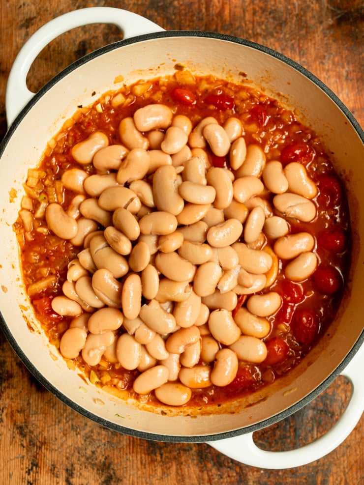Overhead shot of a Dutch oven with drained butter beans just added to a bubbling harissa tomato sauce, not yet stirred in.