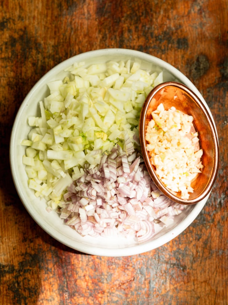 Overhead shot of prepped aromatics — diced fennel and shallots in a white bowl, with minced garlic in a smaller copper bowl alongside — ready to be cooked.