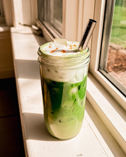 A head-on shot shows an orange cream iced matcha latte with orange cold foam on top. A stainless steel straw sticks out of the glass.