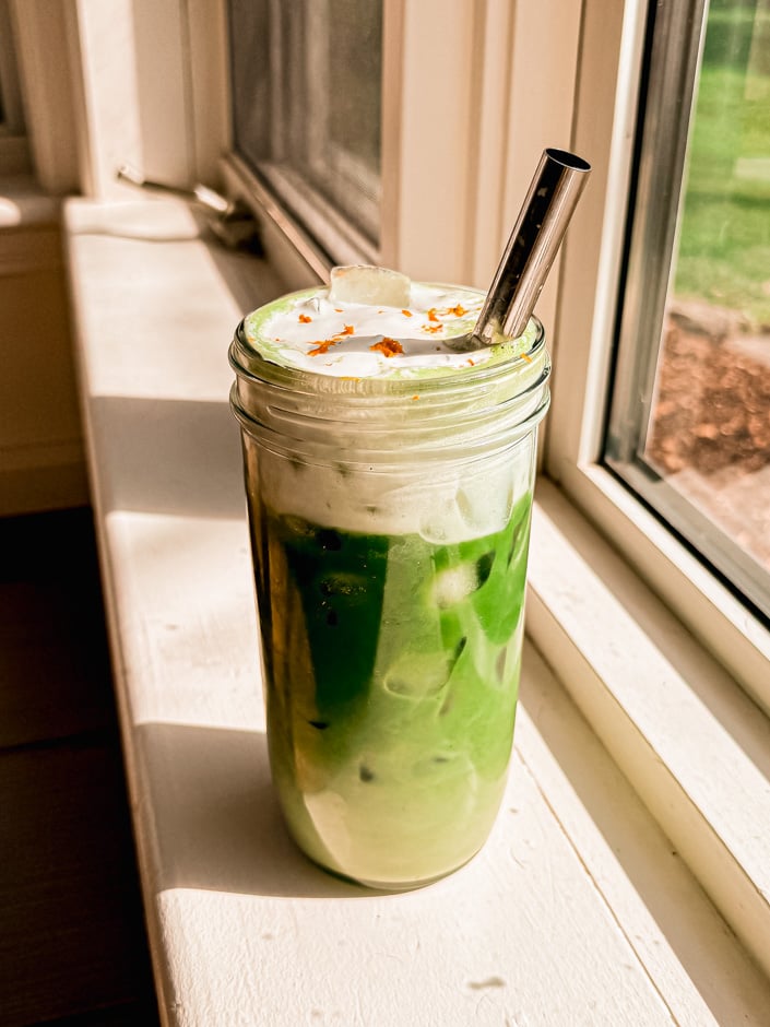 A head-on shot shows an orange cream iced matcha latte with orange cold foam on top. A stainless steel straw sticks out of the glass.
