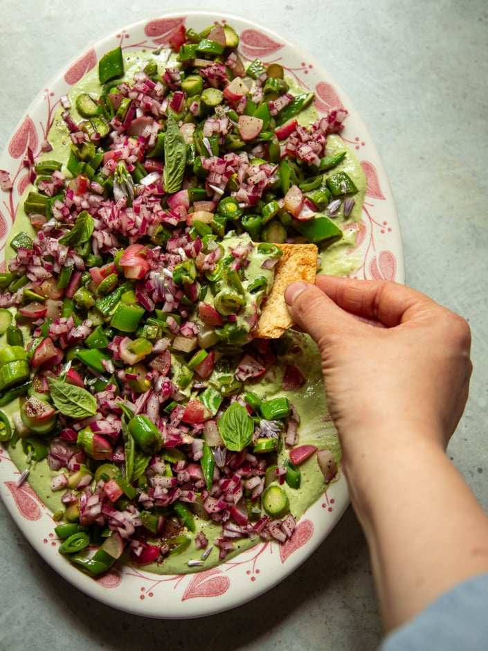 Overhead view of an oval serving platter with a red floral pattern, spread with creamy green cashew goddess sauce and topped with seared spring vegetables. A hand holds a chip, scooping into the dip.