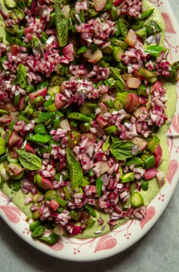 Close-up overhead view of the same platter, showing seared asparagus, snap peas, radishes, and scallions piled over green cashew goddess sauce, garnished with fresh basil leaves and chive blossoms.