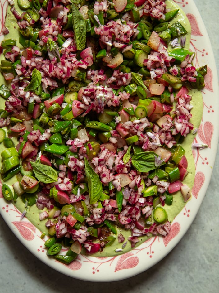 Close-up overhead view of the same platter, showing seared asparagus, snap peas, radishes, and scallions piled over green cashew goddess sauce, garnished with fresh basil leaves and chive blossoms.