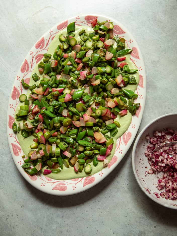Full overhead view of the oval platter with seared spring vegetables over green sauce, with a small bowl of seasoned diced red onion beside it.