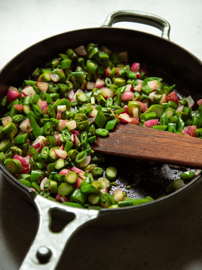 A cast iron skillet filled with chopped spring vegetables — asparagus, snap peas, radishes, and scallions — being stirred with a wooden spatula.