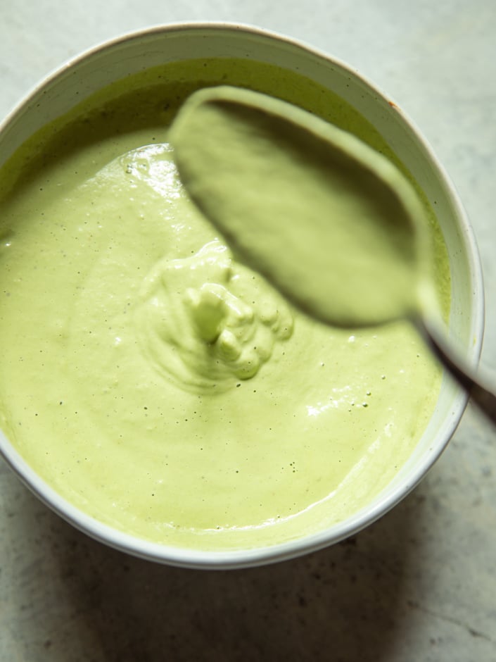 Close-up overhead view of a smooth, pale green cashew goddess sauce in a ceramic bowl, being swirled with a spoon.