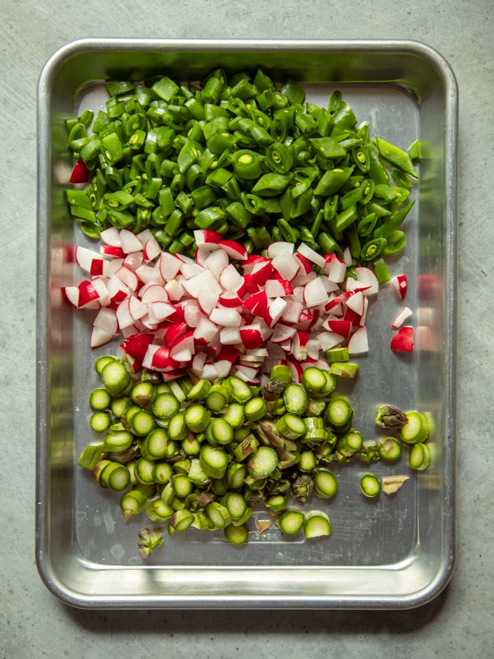 A metal sheet pan with three rows of prepped vegetables: chopped snap peas and scallions at top, diced radishes in the middle, and sliced asparagus at the bottom.