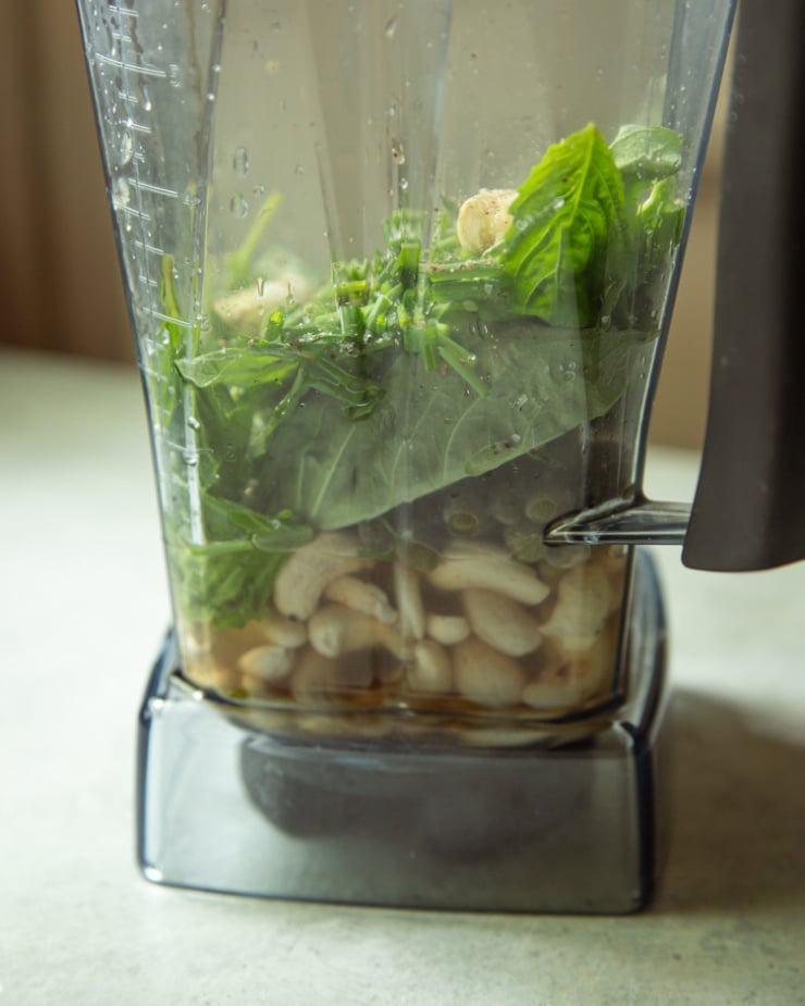 A blender containing cashews, fresh basil, green onions, chives, garlic, water, and other ingredients for the green goddess sauce before blending.