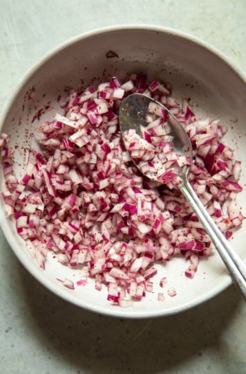 Overhead view of a bowl of finely diced red onion seasoned with sumac and salt, with a spoon resting in it.