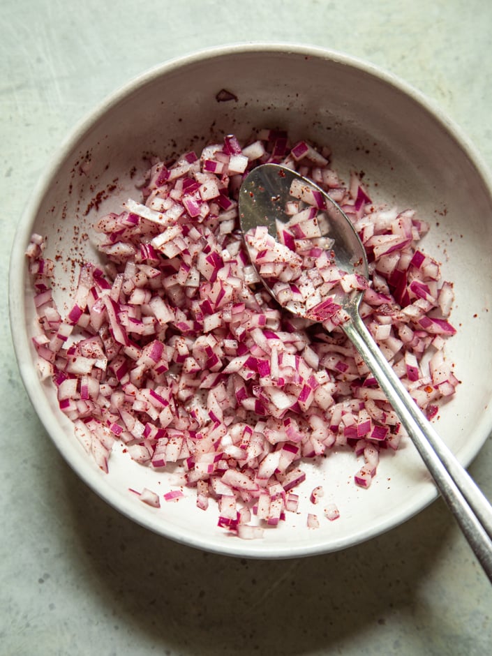 Overhead view of a bowl of finely diced red onion seasoned with sumac and salt, with a spoon resting in it.