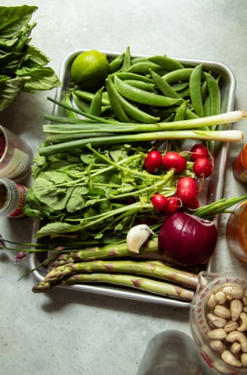 Flat lay of raw ingredients on a metal tray and surrounding surface: snap peas, green onions, radishes, asparagus, red onion, garlic, lime, basil, cashews, sumac, hot sauce, capers, and apple cider vinegar.