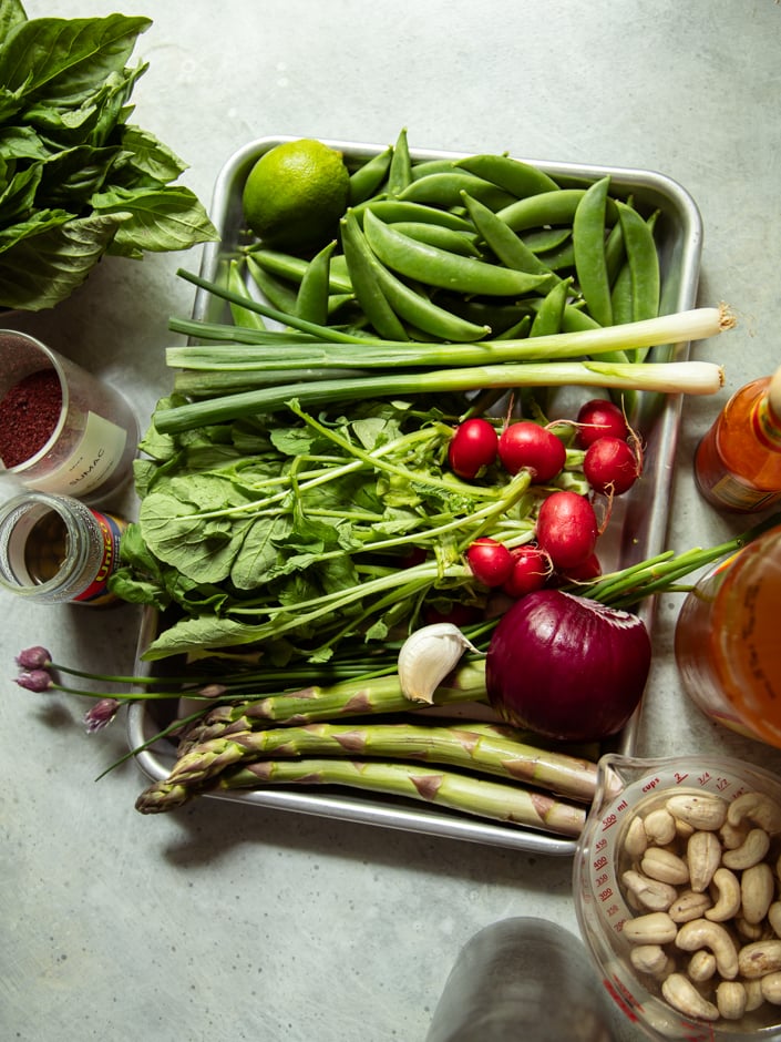 Flat lay of raw ingredients on a metal tray and surrounding surface: snap peas, green onions, radishes, asparagus, red onion, garlic, lime, basil, cashews, sumac, hot sauce, capers, and apple cider vinegar.