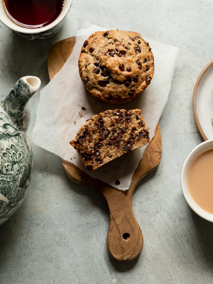 Overhead shot of a whole chocolate chip muffin and a halved muffin on parchment paper on a small wooden cutting board, revealing a dense, chocolate-chip-studded interior, surrounded by a green and white ceramic teapot, a cup of black tea, and a cup of tea with milk.