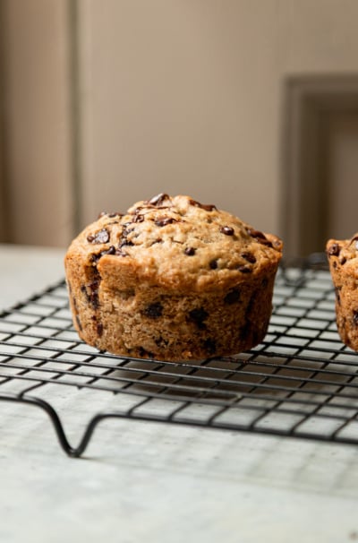 Close-up side view of a large, domed chocolate chip muffin cooling on a black wire rack, with a second muffin partially visible in the background against a warm neutral backdrop.