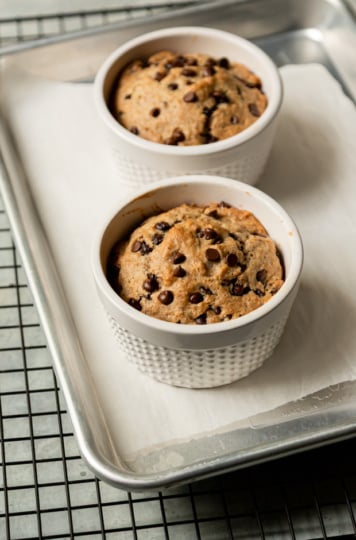 Two freshly baked chocolate chip muffins in white ceramic ramekins on a parchment-lined aluminum baking sheet, domed and golden brown on top.