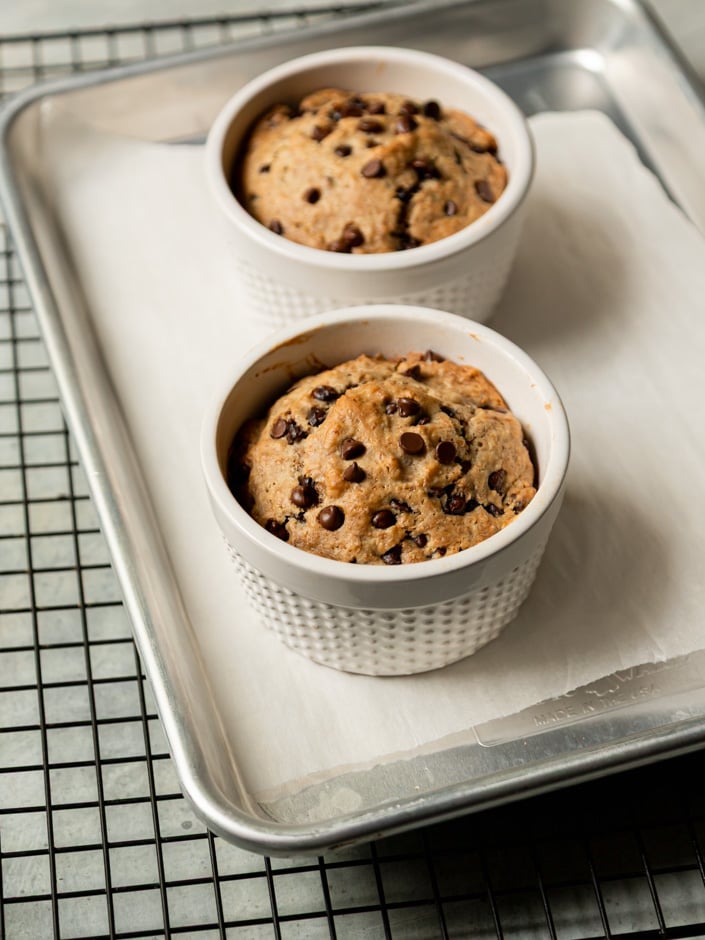 Two freshly baked chocolate chip muffins in white ceramic ramekins on a parchment-lined aluminum baking sheet, domed and golden brown on top.