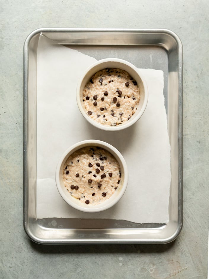 Overhead view of two white ceramic ramekins filled with unbaked chocolate chip muffin batter on a parchment-lined aluminum baking sheet, ready to go in the oven.