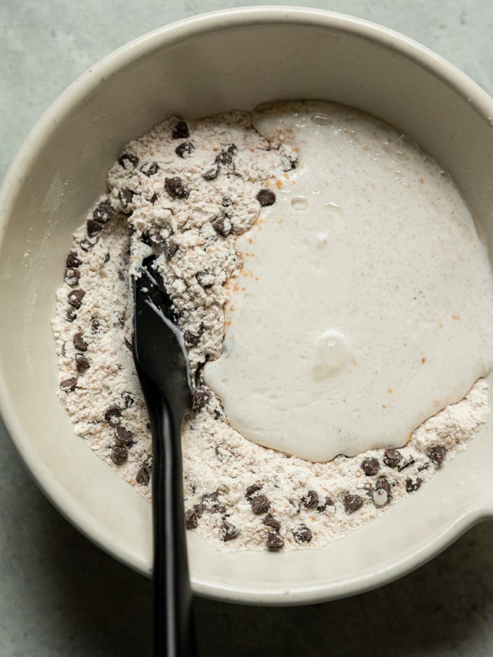 Overhead view of a cream ceramic mixing bowl showing wet ingredients being poured into a dry flour and chocolate chip mixture, with a black spatula beginning to combine them.